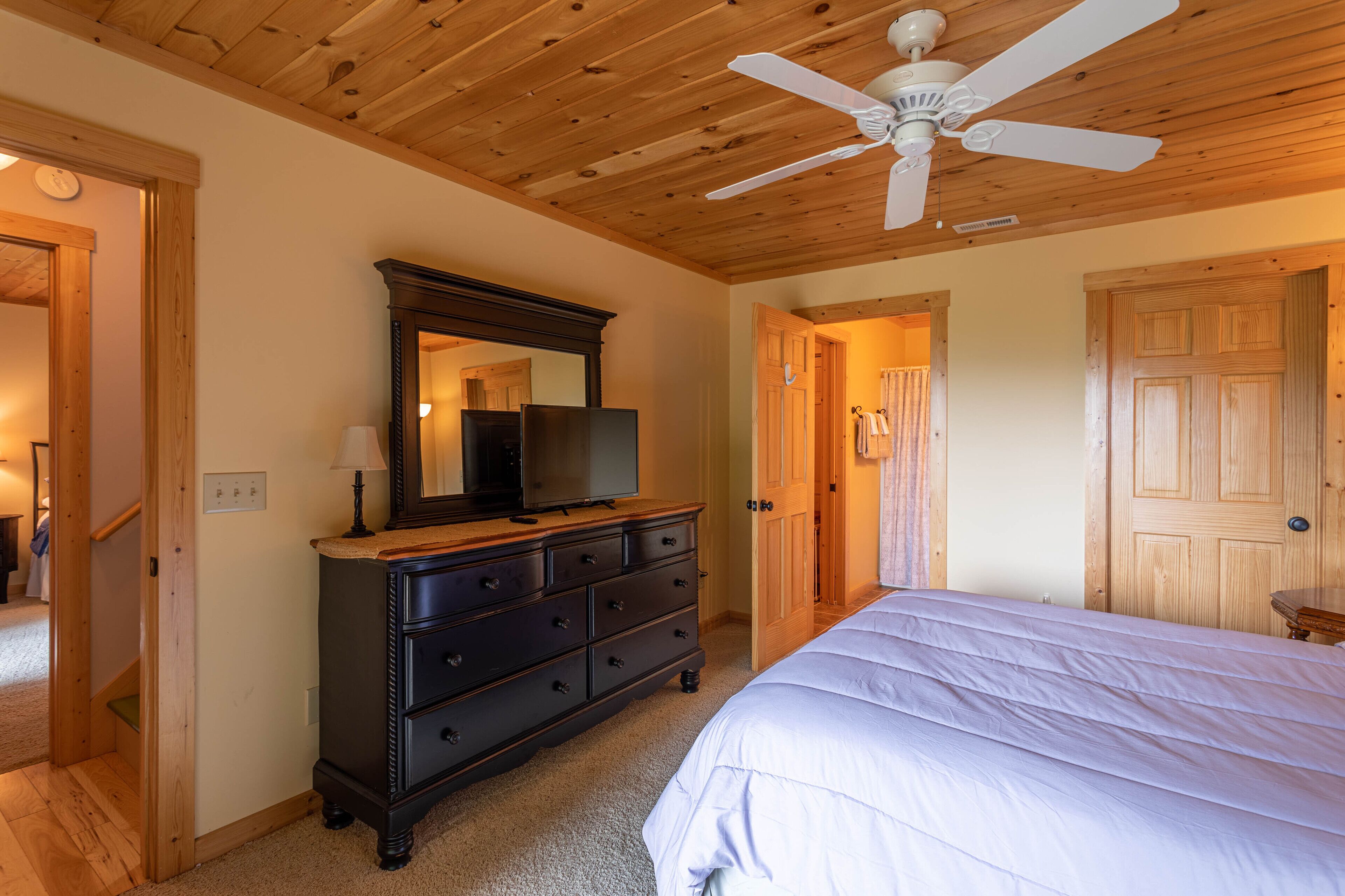 Un dormitorio con techo de madera, un ventilador de techo, una cama, un aparador con espejo y una puerta que da a otra habitación.