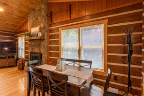 A wooden cabin interior with a dining table, chairs, a fireplace, and a television.