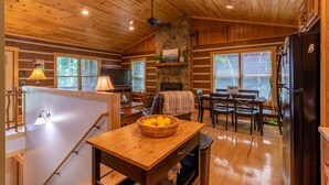 A log cabin interior with a dining area, kitchen, and a stone fireplace. - 1 Relaxing Place - Quaint cottage, great mountain view, hot tub, and fire pit! Private! (Todd)
