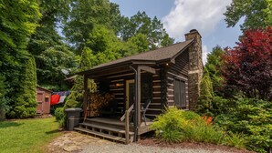 A log cabin with a stone chimney, a porch with steps, and a garden with various plants and a flag. - 1 Relaxing Place - Quaint cottage, great mountain view, hot tub, and fire pit! Private! (Todd)