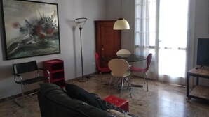 Uma sala de estar com um sofá, uma mesa de jantar com cadeiras, um quadro na parede e uma janela grande.