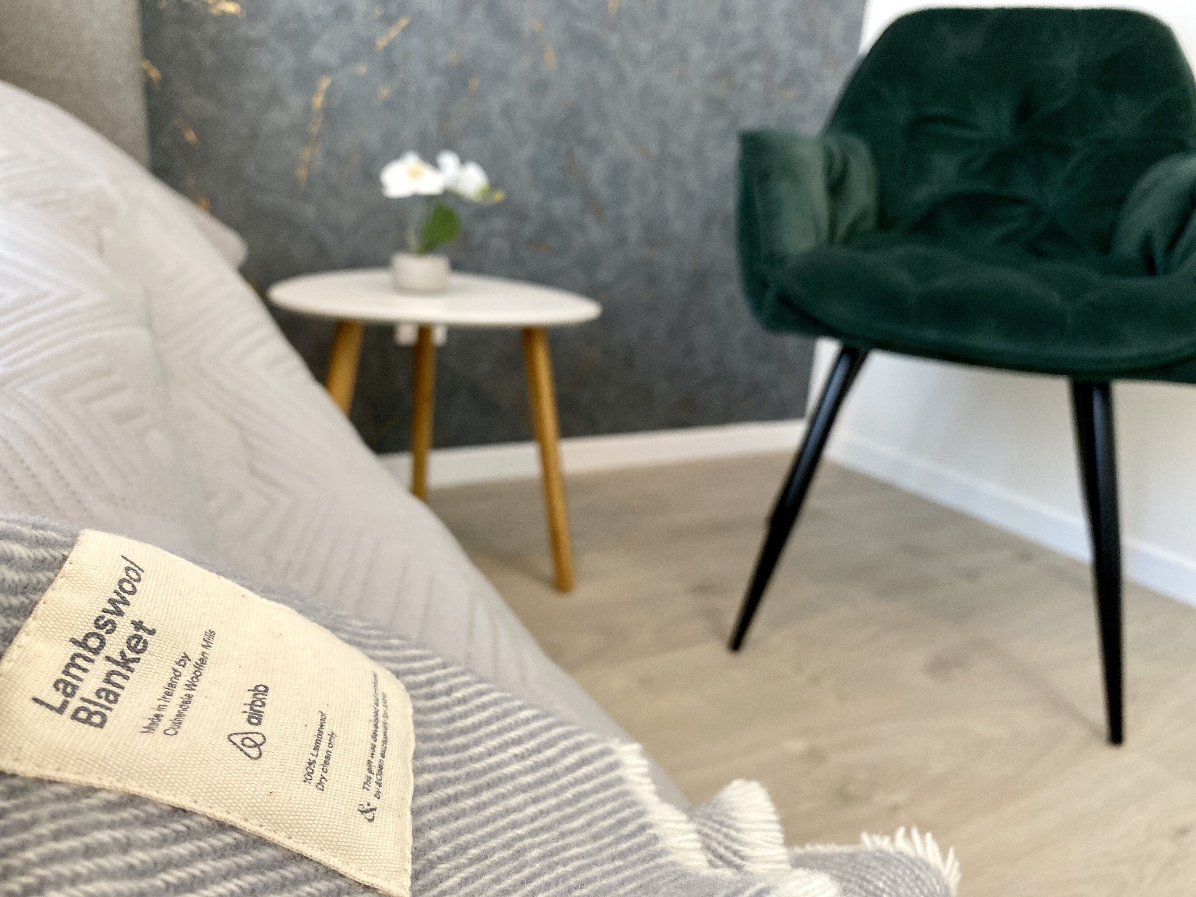 Une chambre avec un lit gris, une chaise verte, une petite table blanche ornée d’une plante et un mur gris à texture.