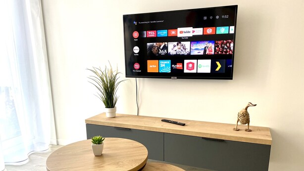 Ein modernes Wohnzimmer mit einem an der Wand befestigten Flachbildfernseher, einem Holzkonsolentisch, zwei runden Holztischen, einer kleinen Topfpflanze und einer dekorativen Vogelfigur.
