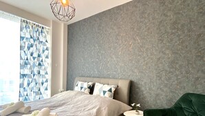 Ein modernes Schlafzimmer mit einem Bett, einem grünen Sessel, einem kleinen Tisch mit einer Pflanze und einem Vorhang mit geometrischem Muster.