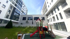 Ein Spielplatz mit Rutsche und Klettergerüsten, umgeben von einer Rasenfläche und einer Bank, mit modernen Wohngebäuden im Hintergrund.