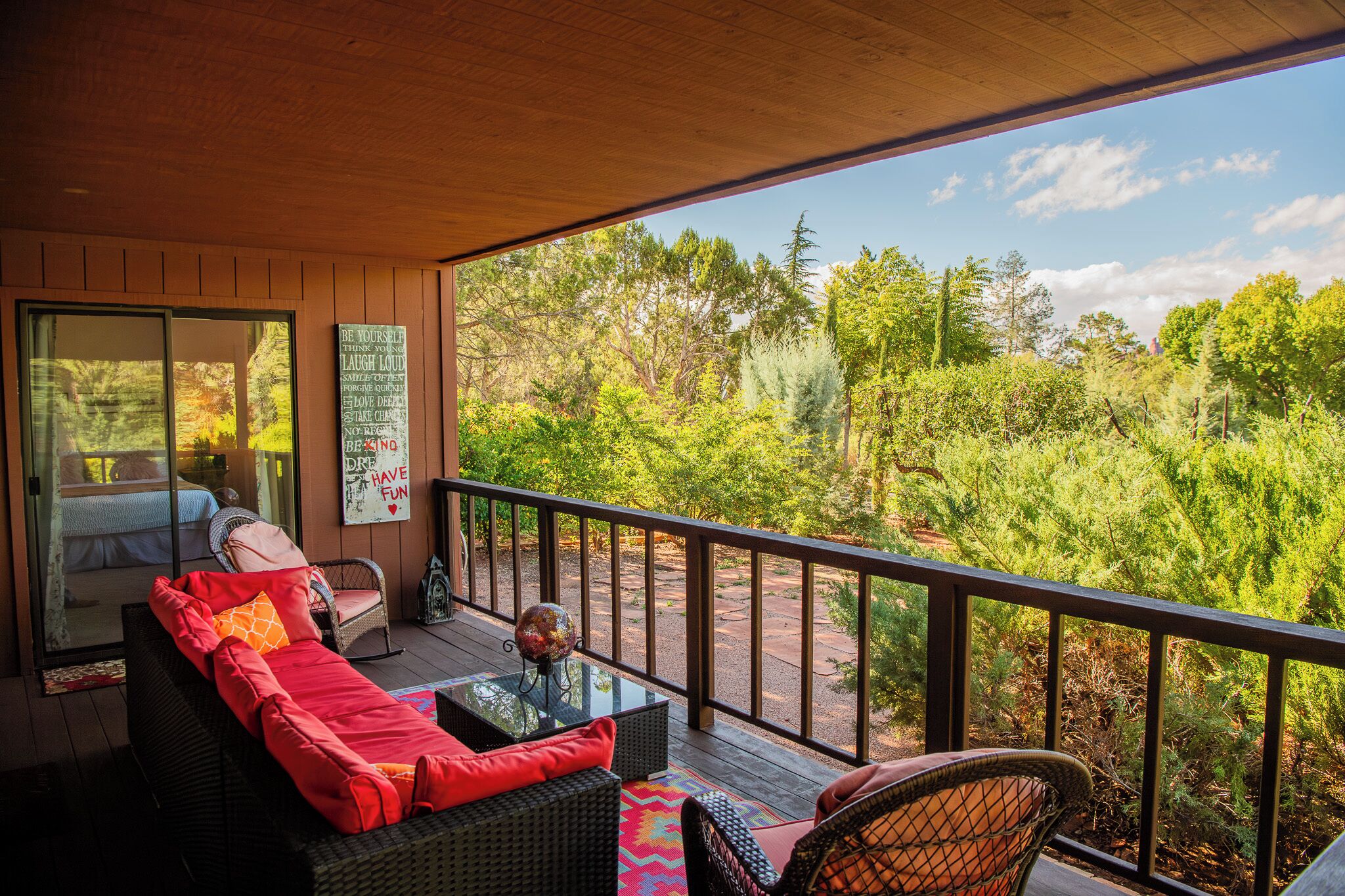 Una terraza techada con mobiliario de mimbre, un ventanal de vidrio que da a un dormitorio y vistas de un área boscosa.