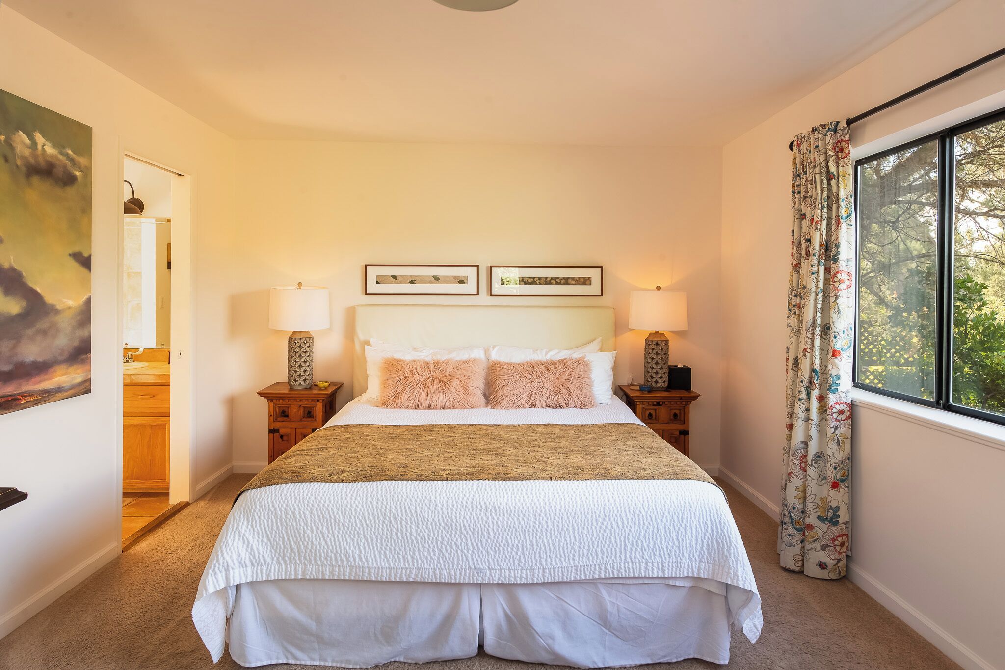 Een net opgemaakt bed met een wit-beige sprei, twee bijpassende kussens en twee bedlampjes. Daarboven hangen twee kunstwerken, en aan de rechterkant een raam met bloemen gordijnen.