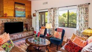 A cozy living room with a stone fireplace, a flat-screen TV, comfortable seating, and a large window with floral curtains. - Luxury Sedona Home with Finnish Sauna and Hot Tub (Sedona)