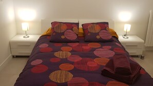 Una cama bien hecha con un edredón estampado, dos lámparas de noche y una manta doblada en el suelo.