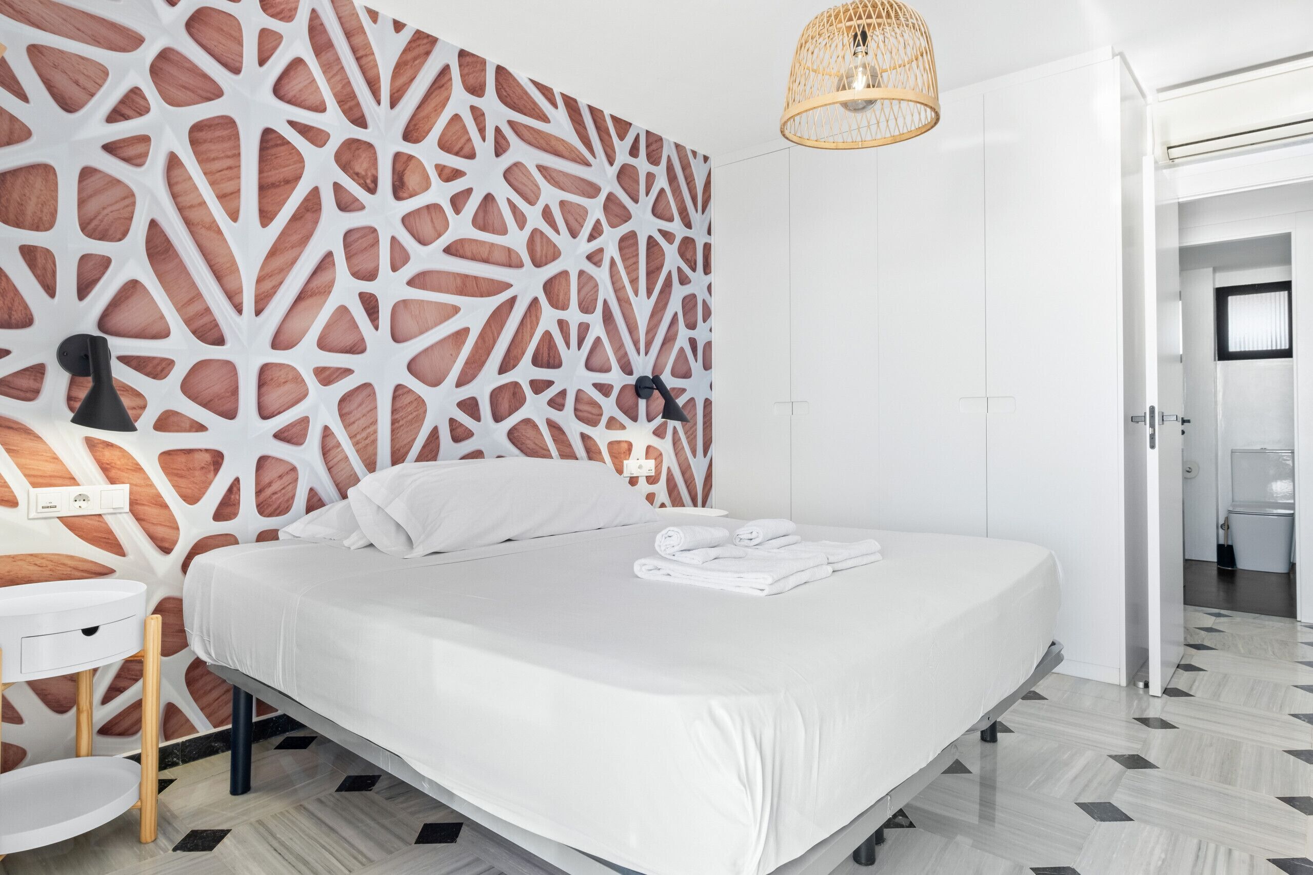 Une chambre moderne avec un mur orné d’un motif géométrique, un lit recouvert de linge de maison blanc, une table de chevet et un luminaire suspendu.