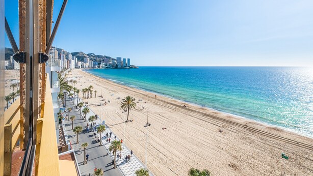 Una playa con cielo azul despejado, una orilla arenosa y un paisaje urbano al fondo.