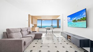 Una sala de estar moderna con un gran ventanal, un sofá modular, una mesa de comedor y un televisor de pantalla plana que muestra una escena de playa.