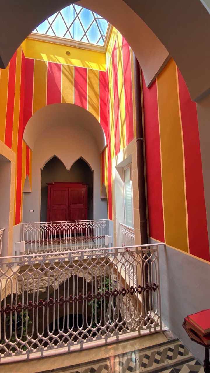 A staircase with a white railing leading to a red door, surrounded by walls with vertical stripes in red, yellow, and orange.