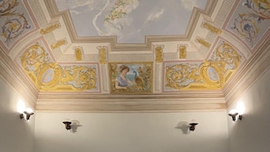 A room with a white door, two white armchairs, a red patterned rug, and a ceiling with ornate gold and blue decorations. - The Musician Angels (Sogliano Cavour)