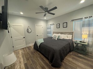 Ein Schlafzimmer mit Bett, Deckenventilator, Nachttisch mit Lampe und Fenster mit Jalousien.