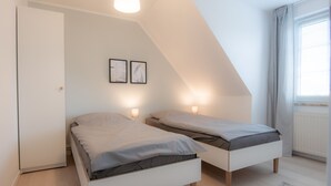Une chambre moderne et bien éclairée, avec deux lits simples, une armoire blanche et une grande fenêtre équipée de stores.