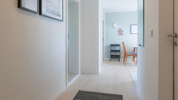 Un couloir avec des tableaux encadrés accrochés au mur, une porte donnant sur une autre pièce et un petit bureau avec une chaise au fond.