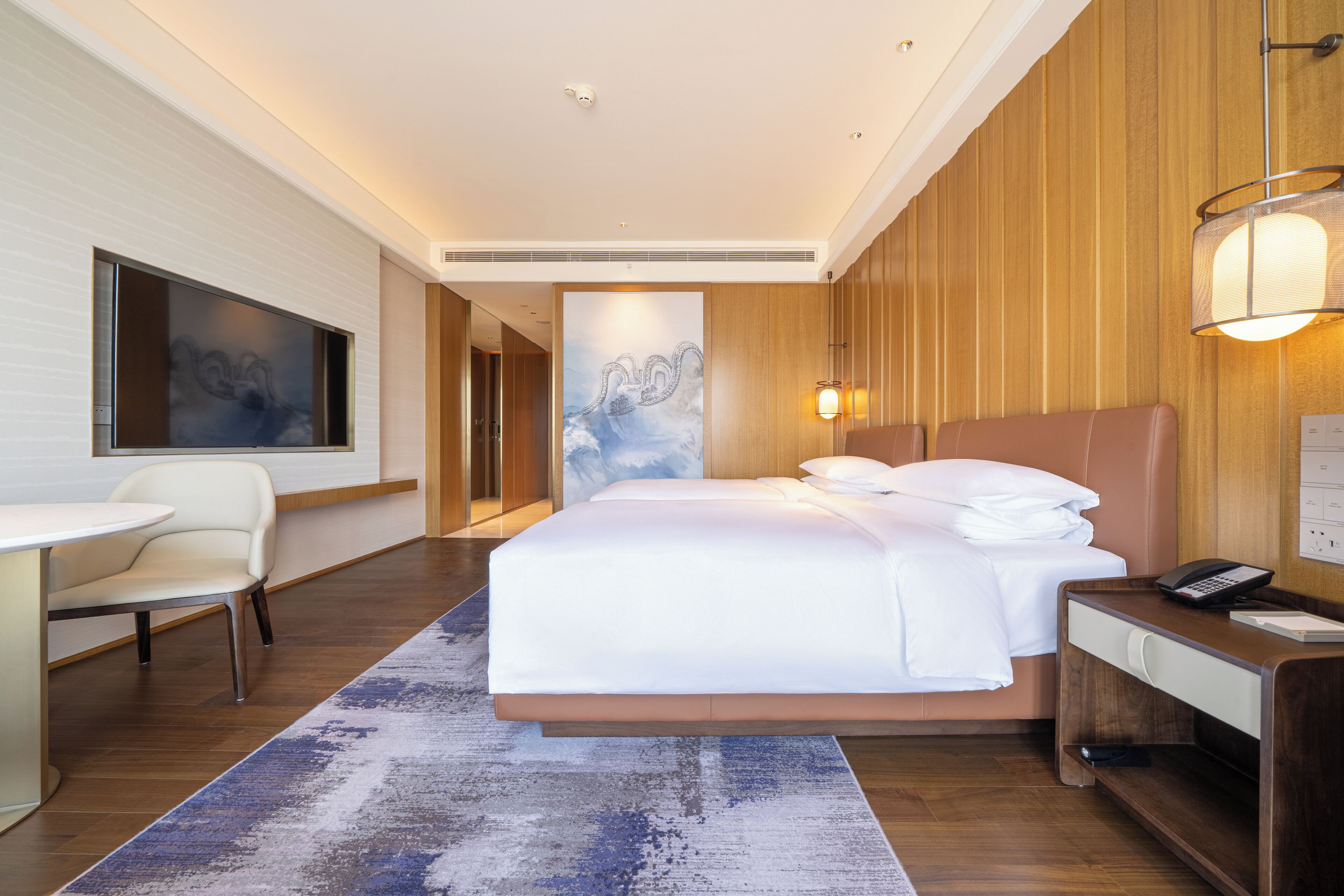A modern hotel room with a large bed, a desk with a telephone, a chair, and a large artwork on the wall.