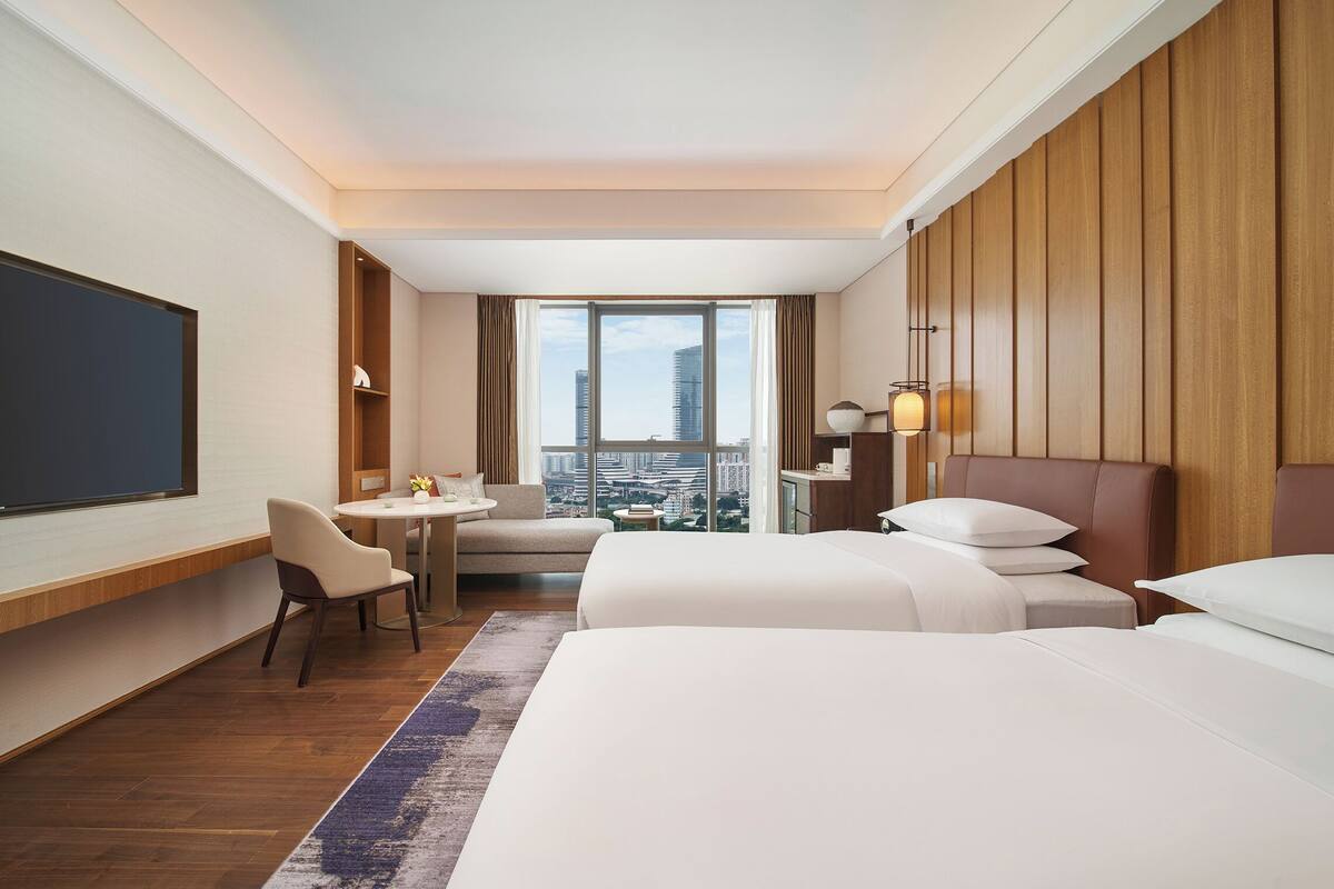Une chambre d’hôtel avec un grand lit, un bureau, une chaise, une petite table et une vue sur la ville par une grande fenêtre.
