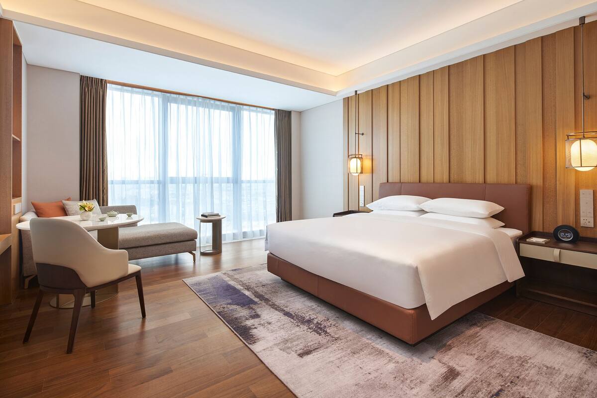 Une chambre d’hôtel moderne dotée d’un grand lit, d’un coin salon avec une chaise et une table, et d’une grande fenêtre avec des rideaux.