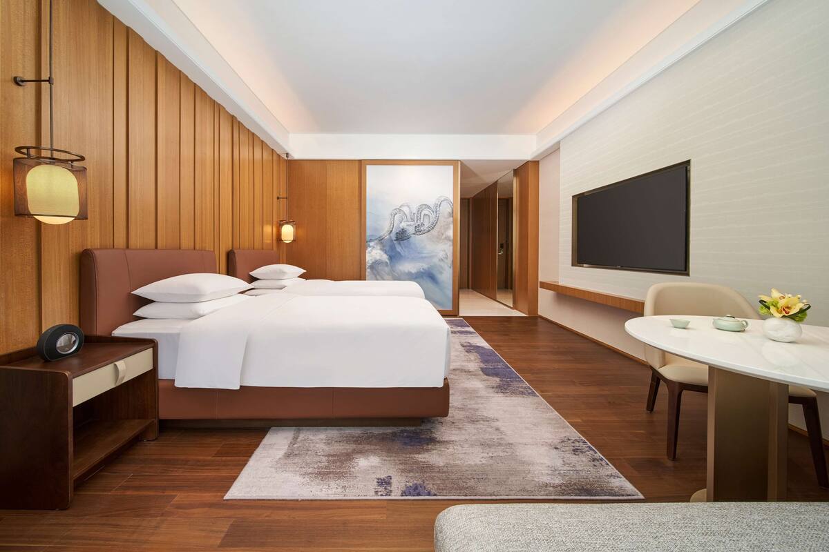 Une chambre d’hôtel moderne avec un mur lambrissé en bois, un grand lit avec du linge de lit blanc, un bureau avec une lampe, un téléviseur à écran plat et une petite table à manger agrémentée d’un vase de fleurs.