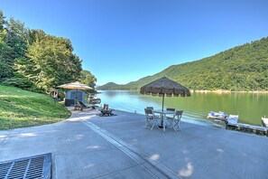 Un patio junto al lago con un toldo de paja, asientos y un pequeño edificio, probablemente un baño.
