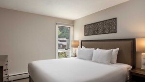 A hotel room with a large bed, a nightstand with a lamp, a wall-mounted artwork, and a window with a view of buildings and trees. - Vail 21 407 - 2 Bedroom Diamond (Vail)
