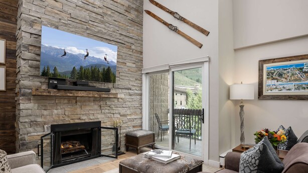 A living room with a stone fireplace, a television mounted on the wall, and a large window overlooking a deck. - Vail 21 407 - 2 Bedroom Diamond (Vail)