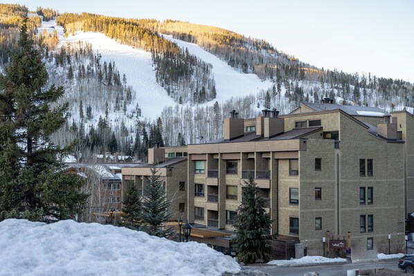 A multi-story building with a brick facade, likely a hotel or apartment complex, surrounded by snow-covered trees and a mountain in the background. - Vail 21 407 - 2 Bedroom Diamond (Vail)