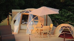 Une structure en forme de dôme géodésique, comprenant une table et des chaises, entourée d’une terrasse et d’arbres.