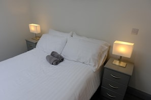 Una cama bien hecha con sábanas blancas y dos toallas enrolladas, flanqueada por dos mesitas de noche con lámparas, en una habitación sencilla con una pared lisa y un cuadro de interruptores.