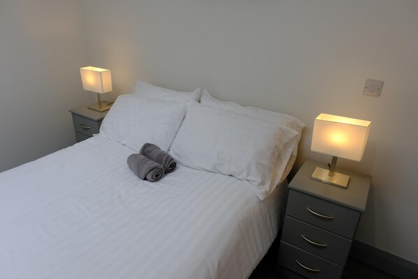 Una cama bien hecha con sábanas blancas y dos toallas enrolladas, flanqueada por dos mesitas de noche con lámparas, en una habitación sencilla con una pared lisa y un cuadro de interruptores.