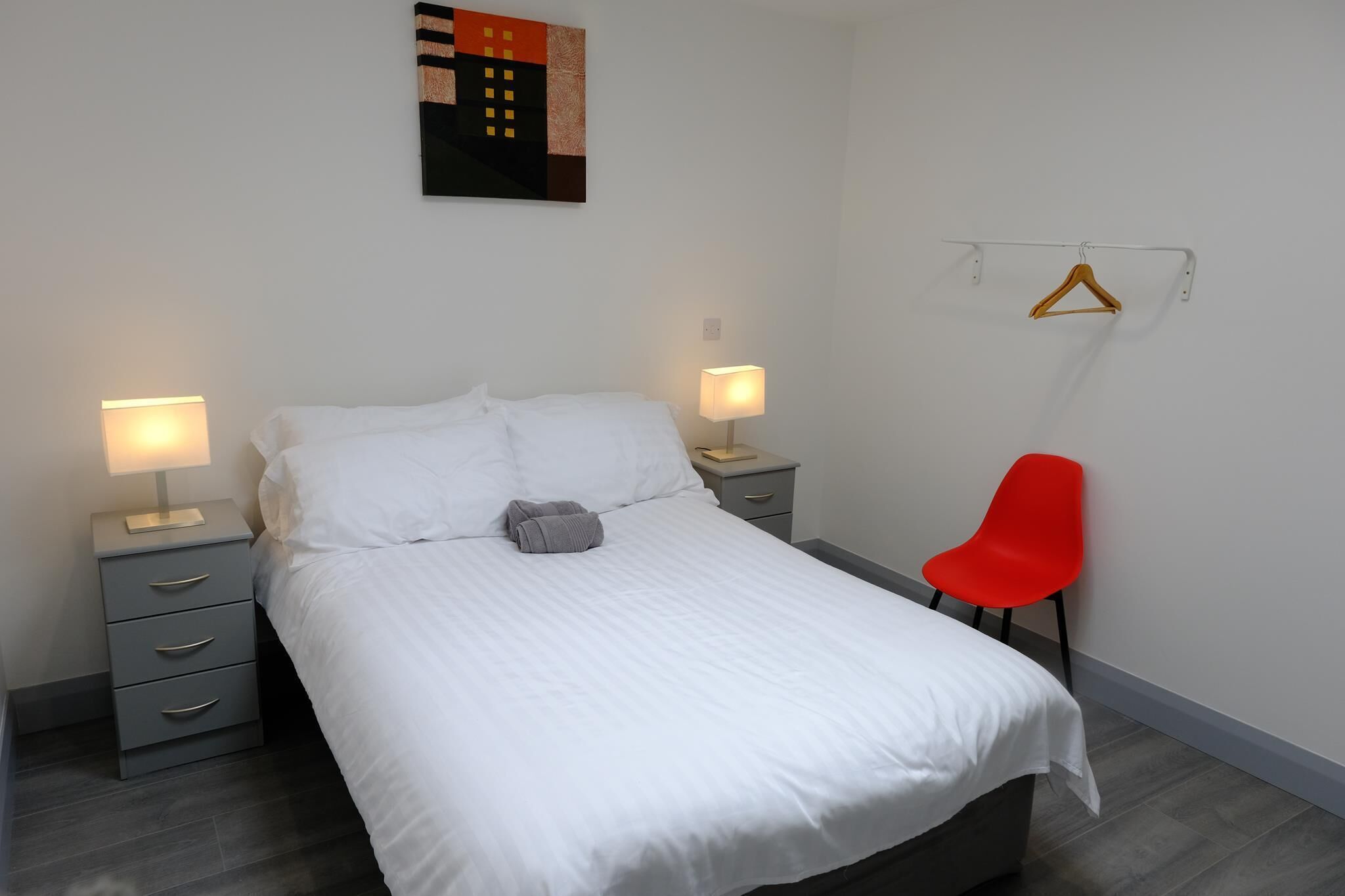 A neatly made bed with white linens, two bedside tables with lamps, a grey dresser, and a red chair.