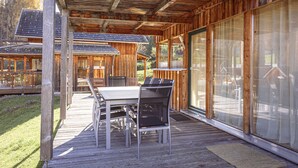 Een houten terras met een tafel en stoelen, grote schuifpuien en uitzicht op een bebost gebied.