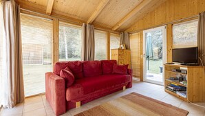 Un salón acogedor con un sofá rojo, techo de madera, ventanas amplias con persianas, un televisor y una estantería.