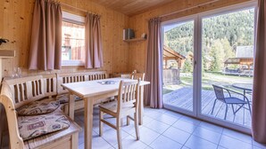 Interior de una cabaña de madera con una mesa de comedor, sillas, un banco acolchado y un ventanal corredizo que da a una terraza exterior.