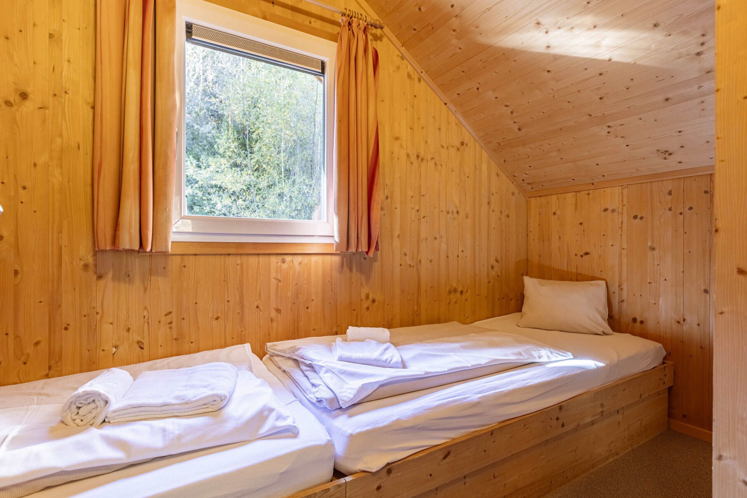 Ein kleines, hölzernes Zimmer mit einem Einzelbett, einem Fenster mit Vorhängen und Blick auf Bäume.