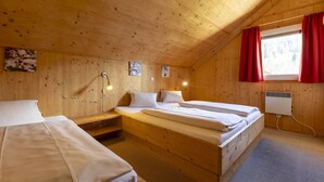 A wooden cabin room with two beds, a window with red curtains, and a wall-mounted heater.