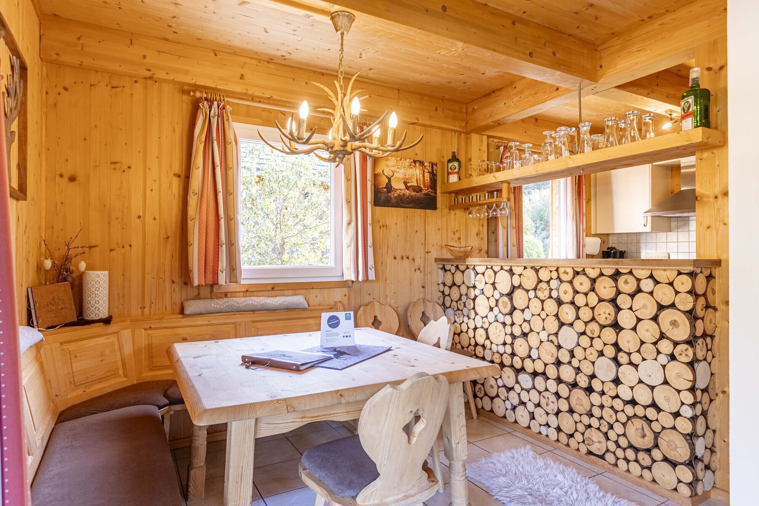 A wooden cabin interior with a dining table, bench, chandelier, and a wall of stacked logs.