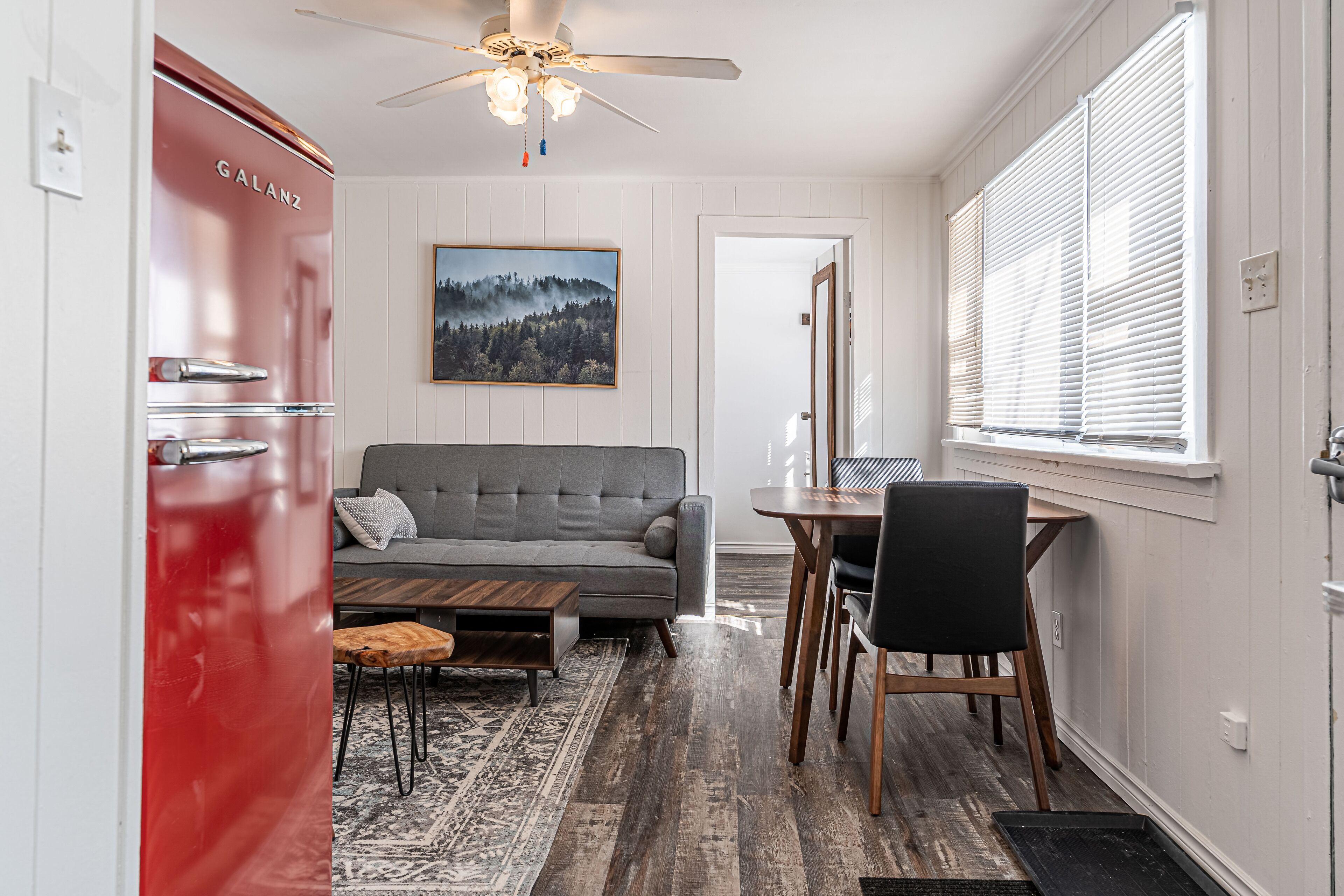A modern living room with a grey sofa, wooden coffee table, and a dining area with a table and chairs. A red appliance is visible on the left, and a painting hangs on the wall.