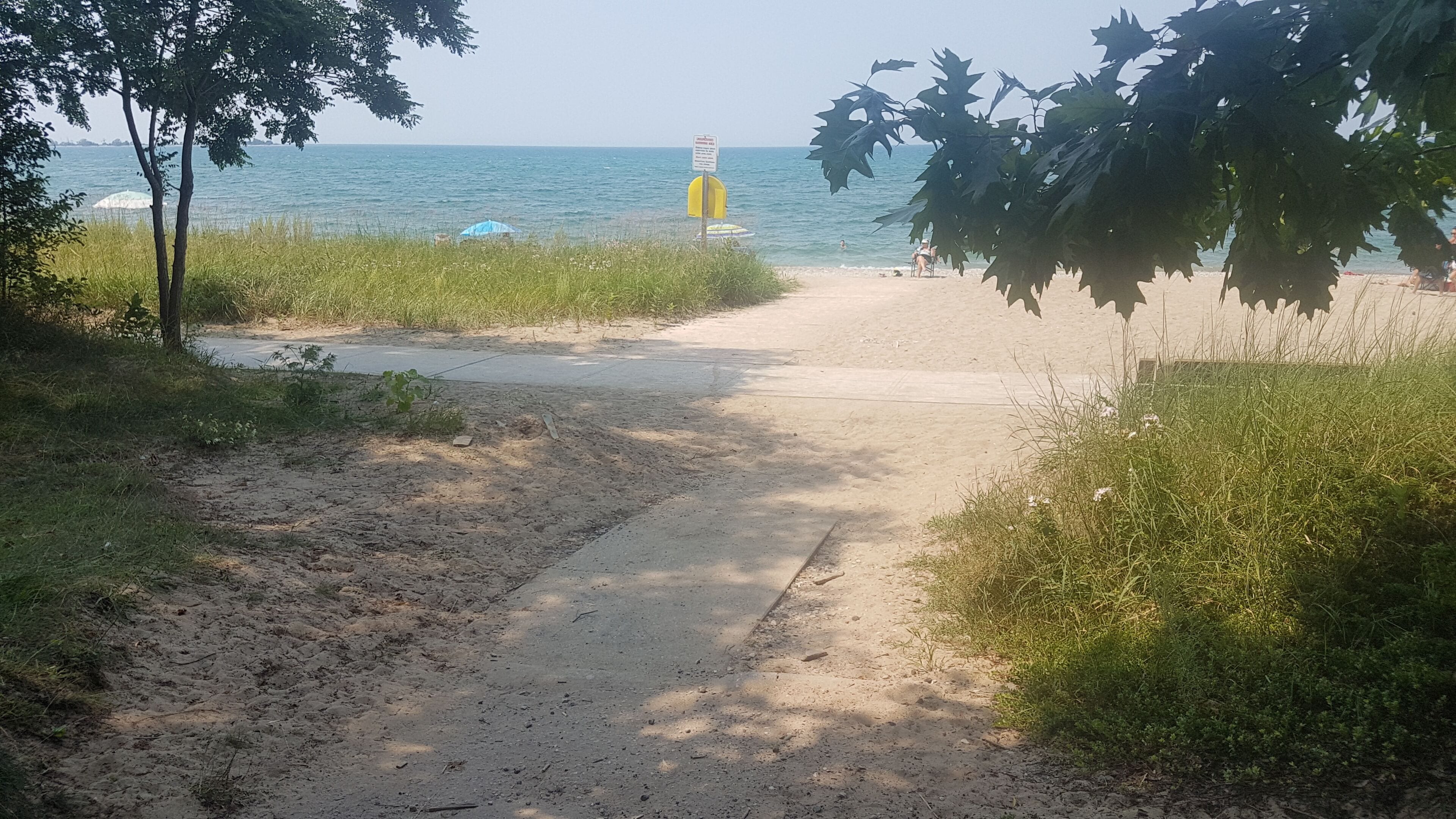Un sentier sablonneux mène à une plage, avec un panneau indicateur et un poste de surveillance des sauveteurs visible au loin.