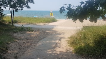 Un sendero arenoso que lleva a una playa, con un cartel indicador y una torre de salvavidas a lo lejos.