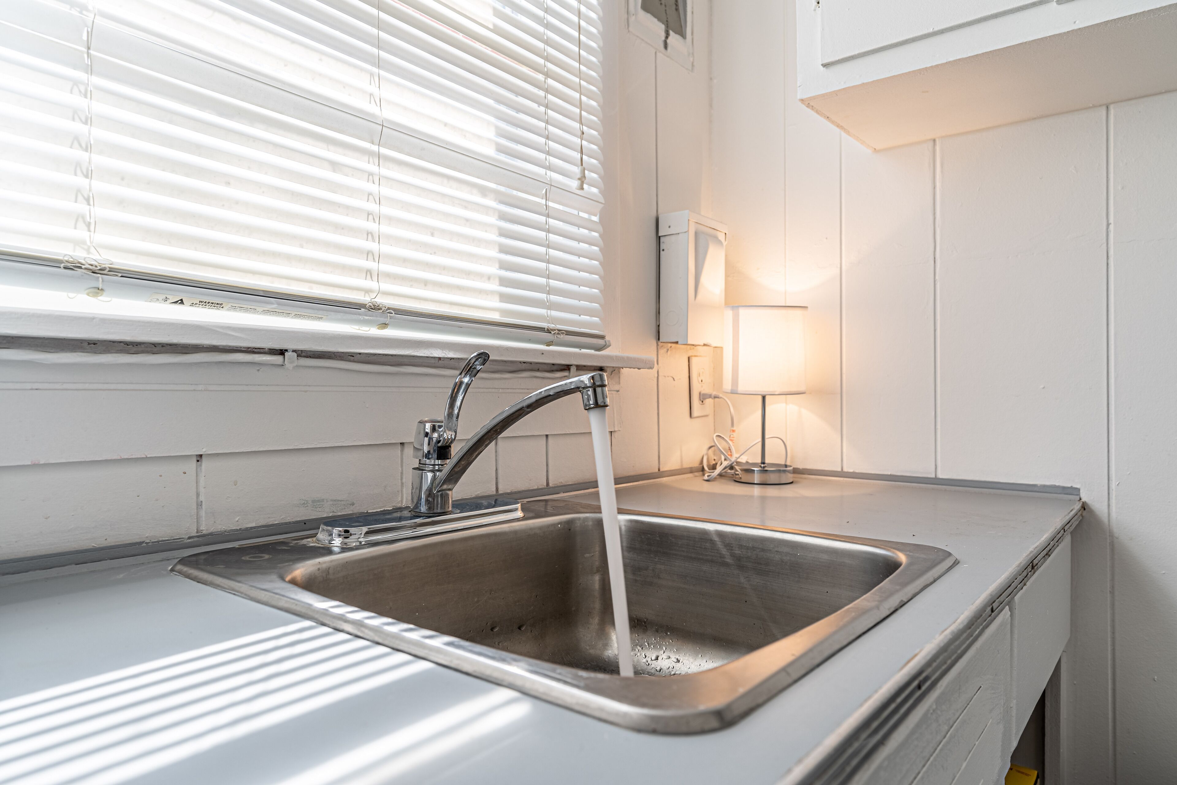 A stainless steel kitchen sink with running water, a window with blinds, and a wall-mounted light fixture.