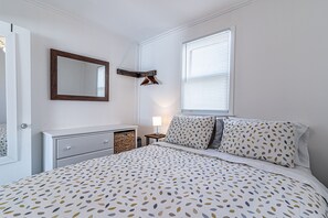 A bedroom with a bed, a dresser, a mirror, a nightstand with a lamp, and a window with blinds.