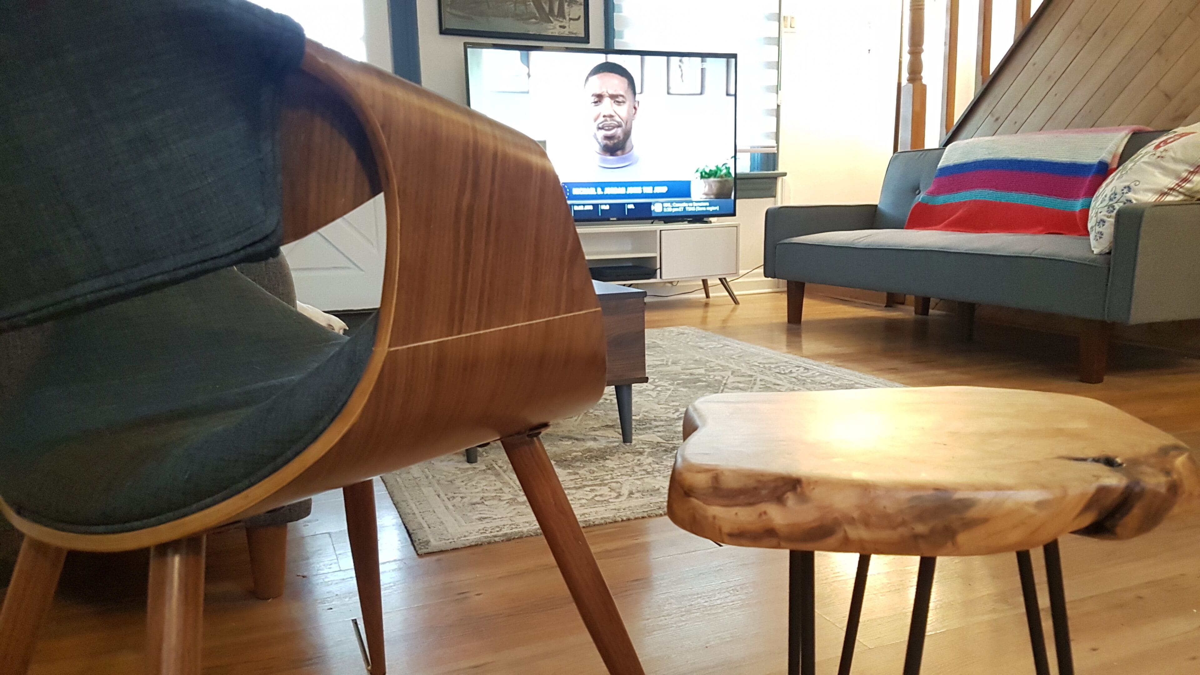 A modern living room with a wooden chair, a small wooden stool, a grey sofa, and a television displaying a news program.