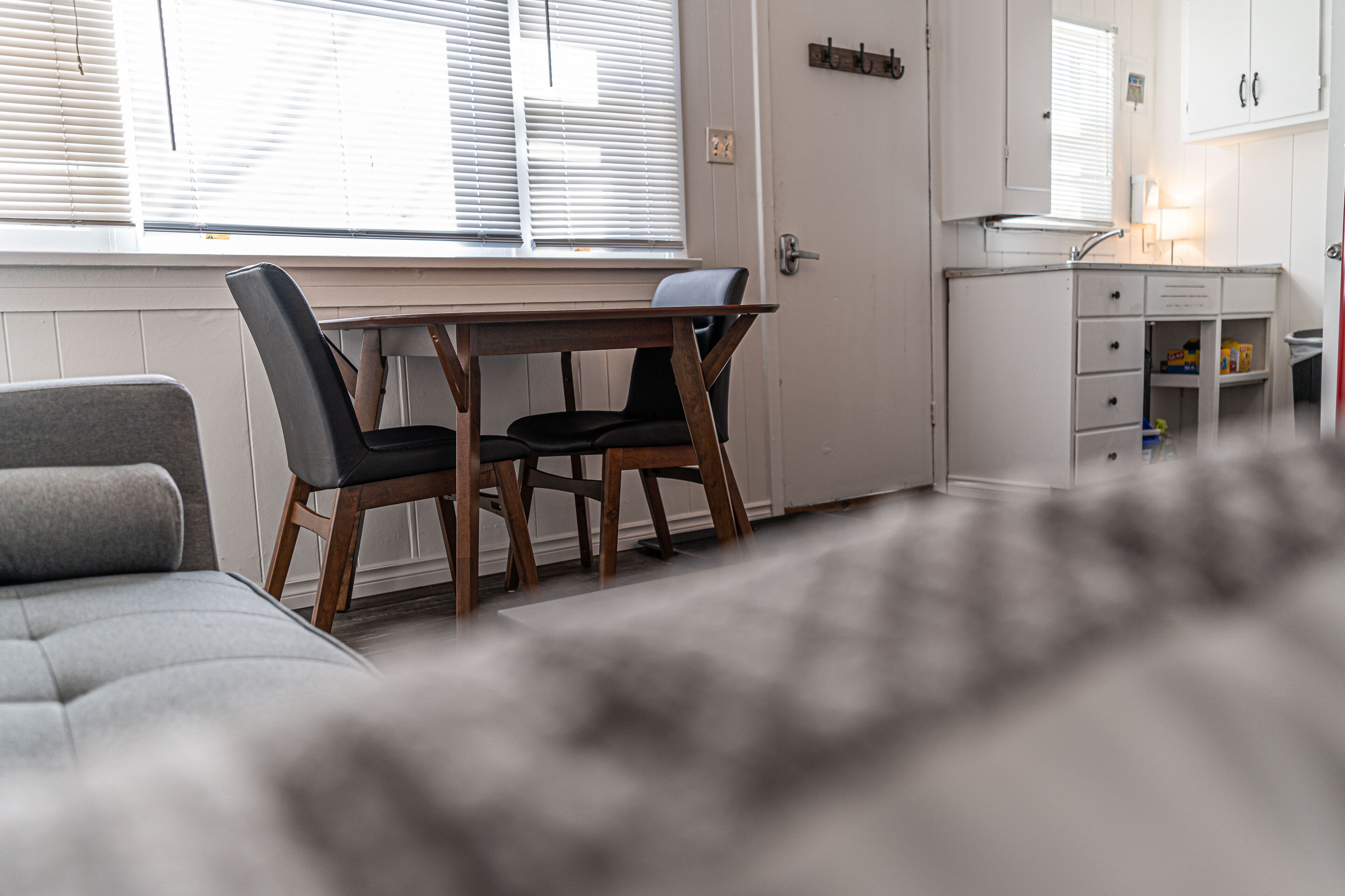 A room with a dining table and chairs, a sofa, a small kitchenette, and a window with blinds.