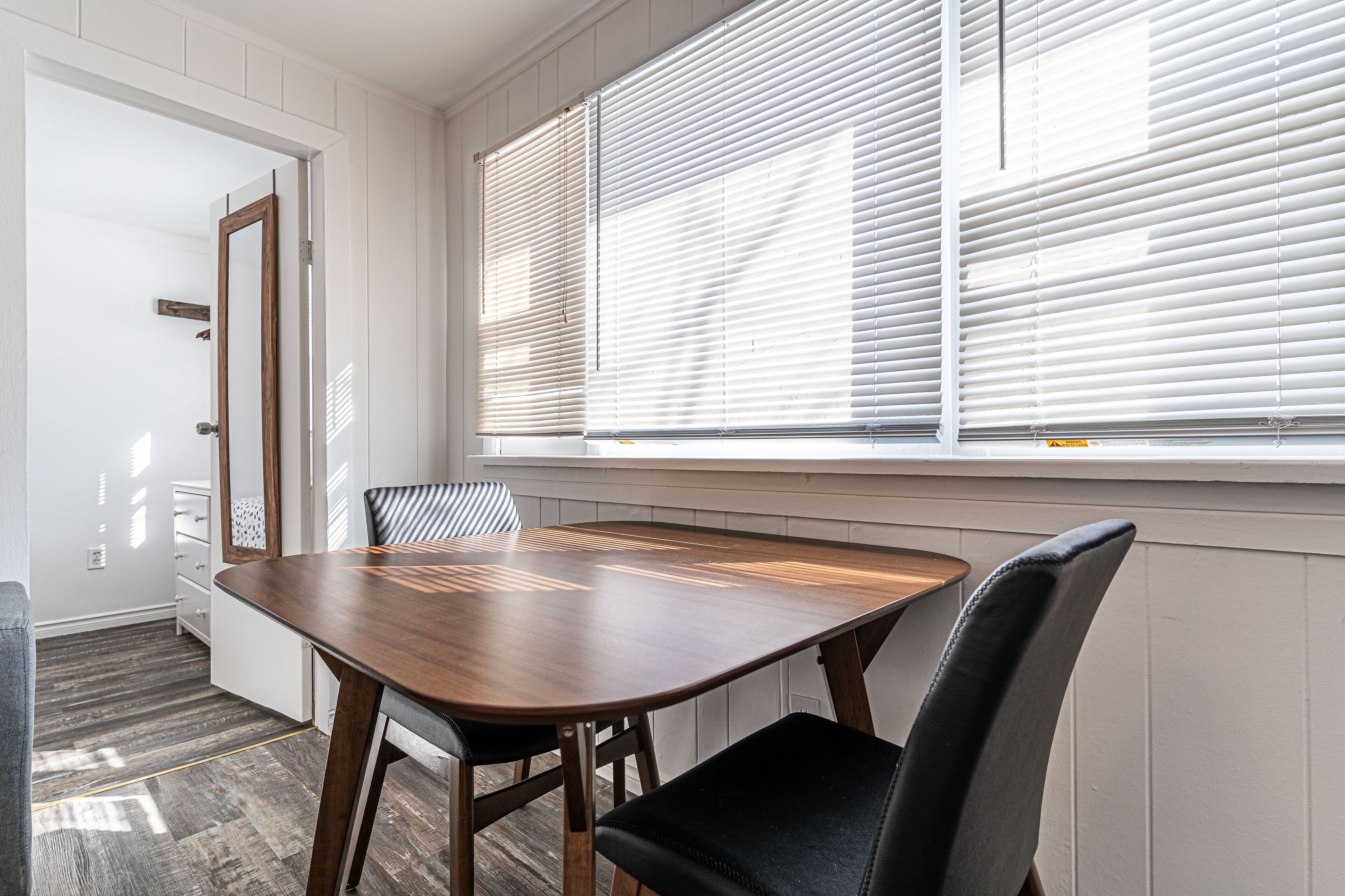 A dining area with a wooden table and chairs, a window with blinds, and a door leading to another room.