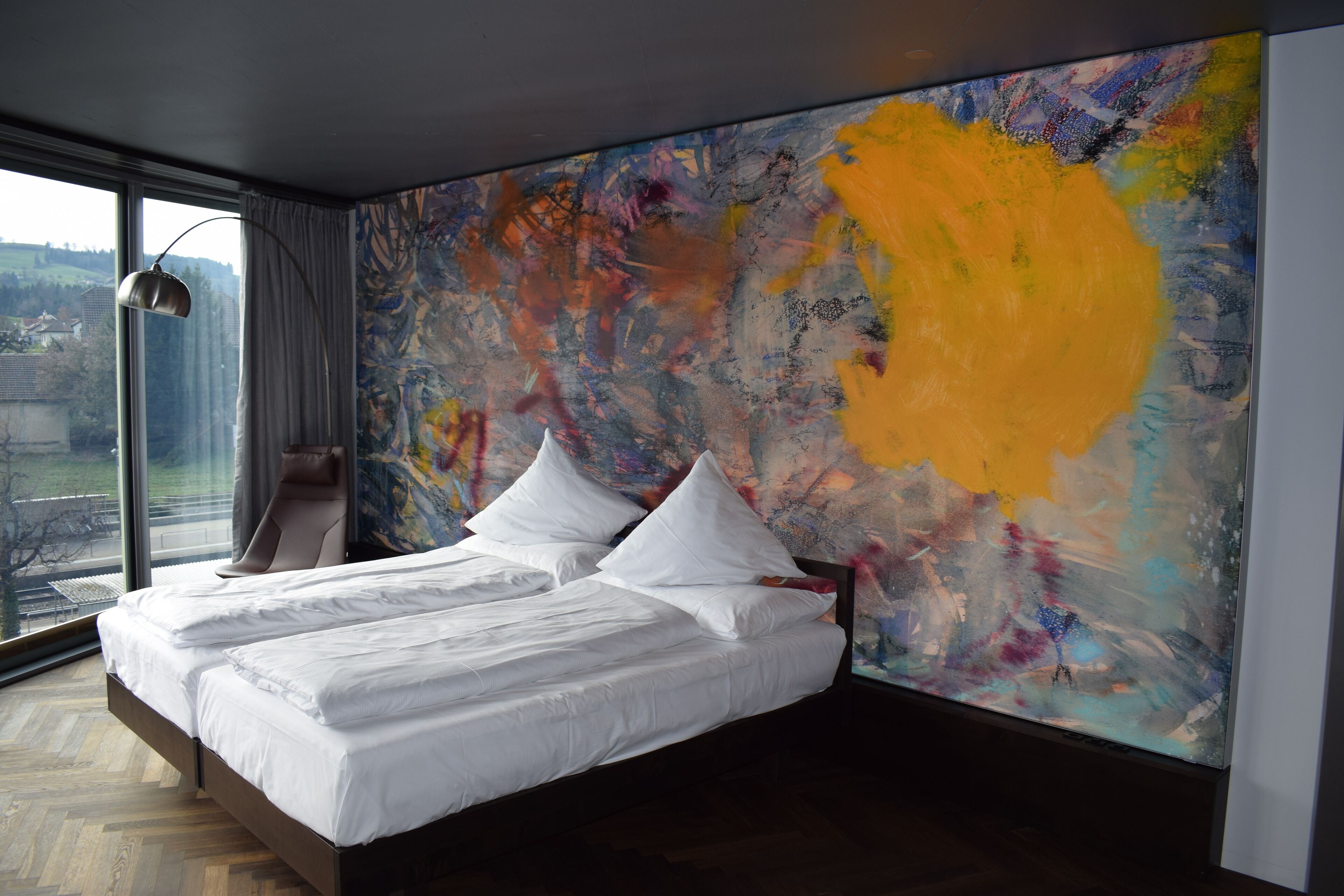 A modern bedroom with a large bed, a view of the city, and a colorful abstract painting on the wall.