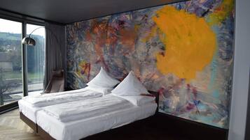 A modern bedroom with a large bed, a view of the city, and a colorful abstract painting on the wall.