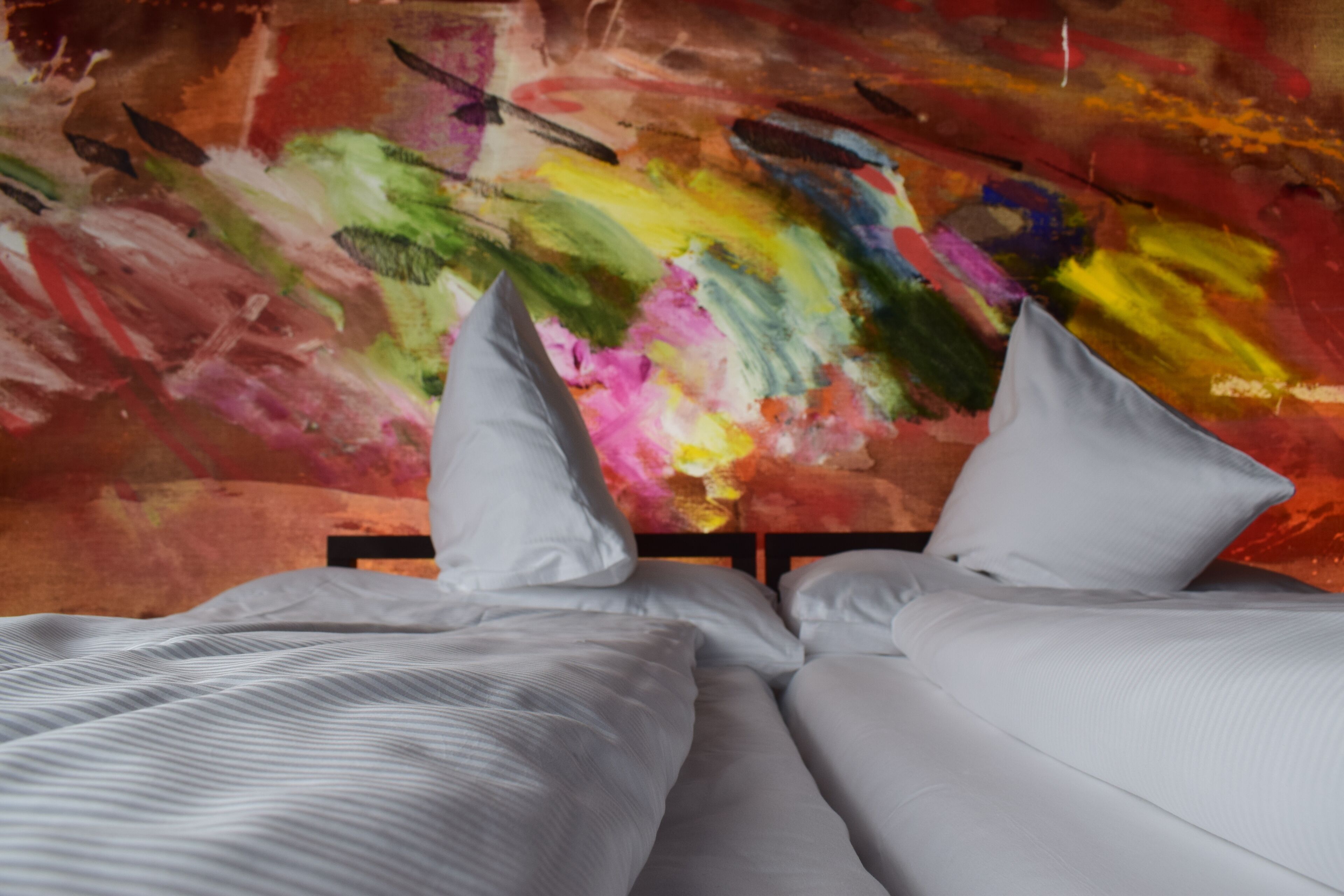Un lit avec du linge de lit blanc et des oreillers, placé devant un tableau abstrait aux couleurs vives accroché au mur.